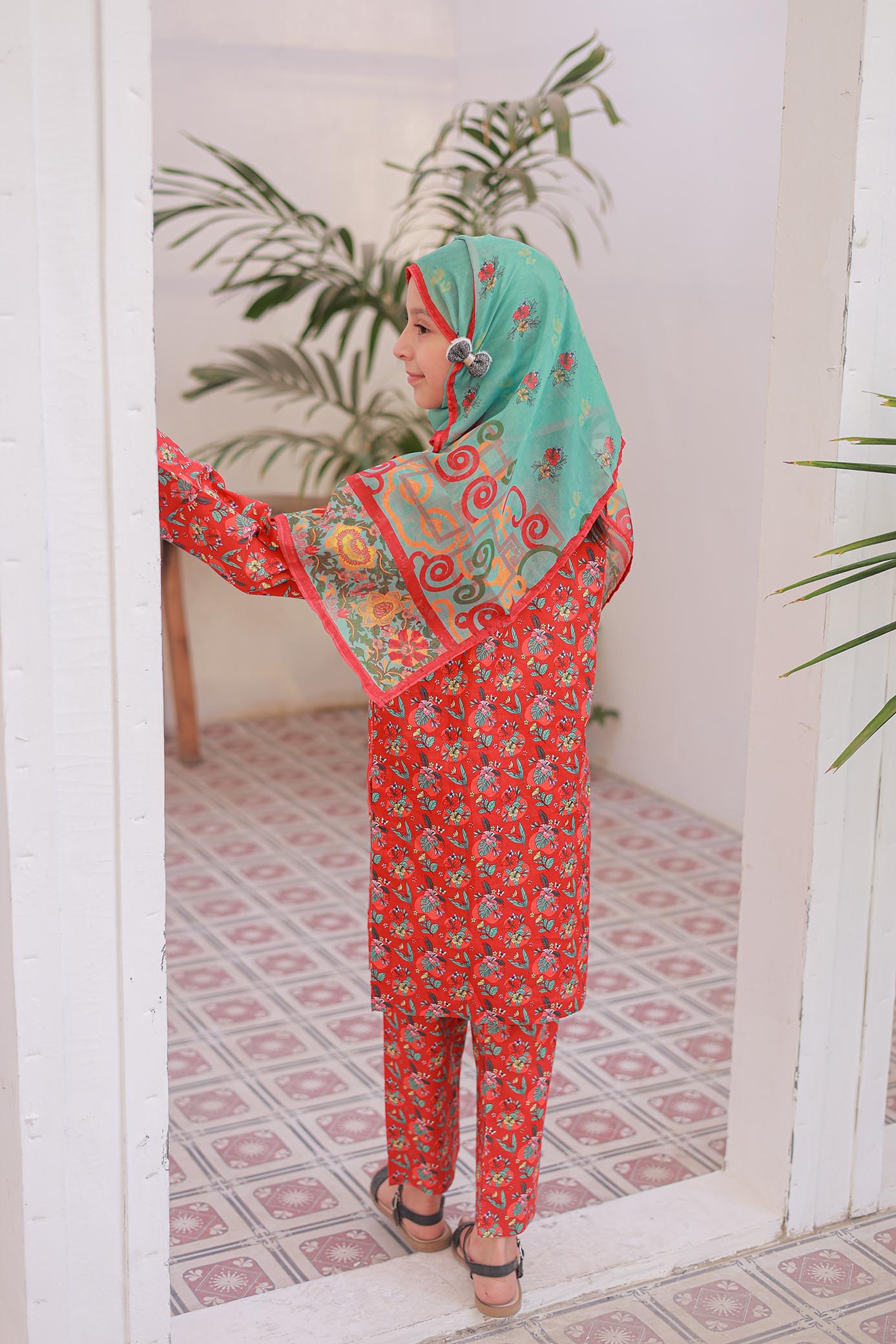 Woman wearing a red and green traditional outfit with a patterned headscarf in a room with plants.