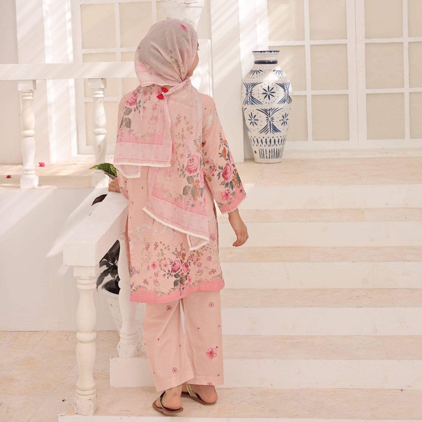 Woman in a pink traditional outfit standing in a decorated room with a vase and plants.