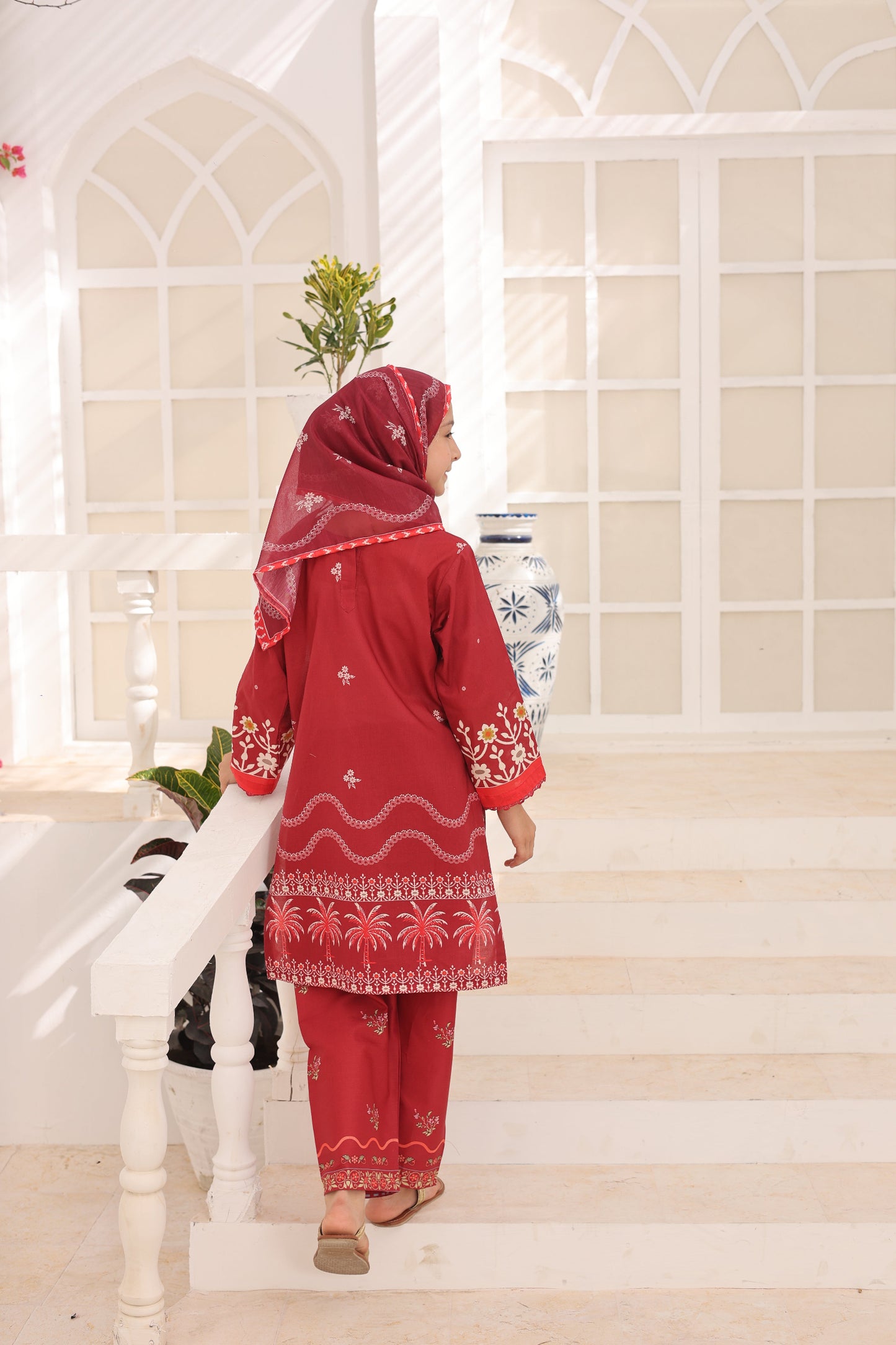 Woman in a red traditional outfit standing on a staircase with a decorative wall in the background.
