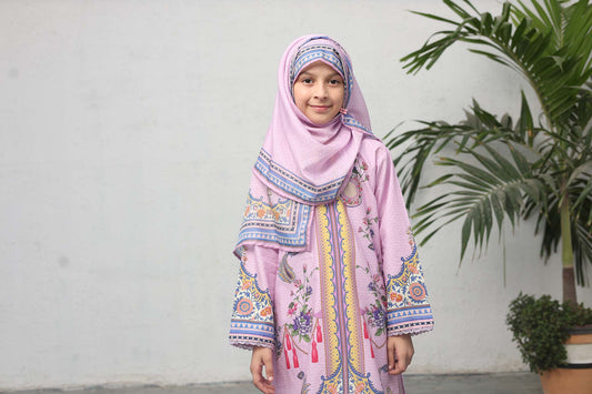 Young girl wearing a pink traditional outfit with a matching hijab, standing against a plain background with a plant.