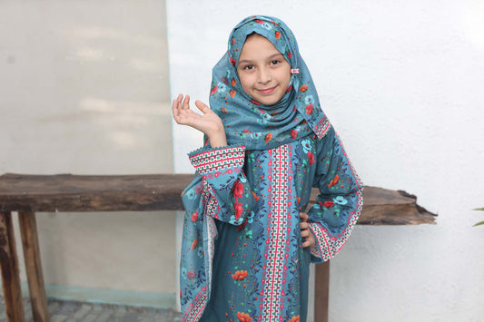 Young girl wearing a blue traditional outfit with a matching hijab, standing in front of a wooden table.