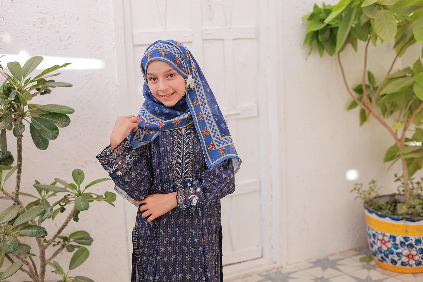 Child wearing a blue and white patterned hijab and dress, standing indoors with plants in the background.