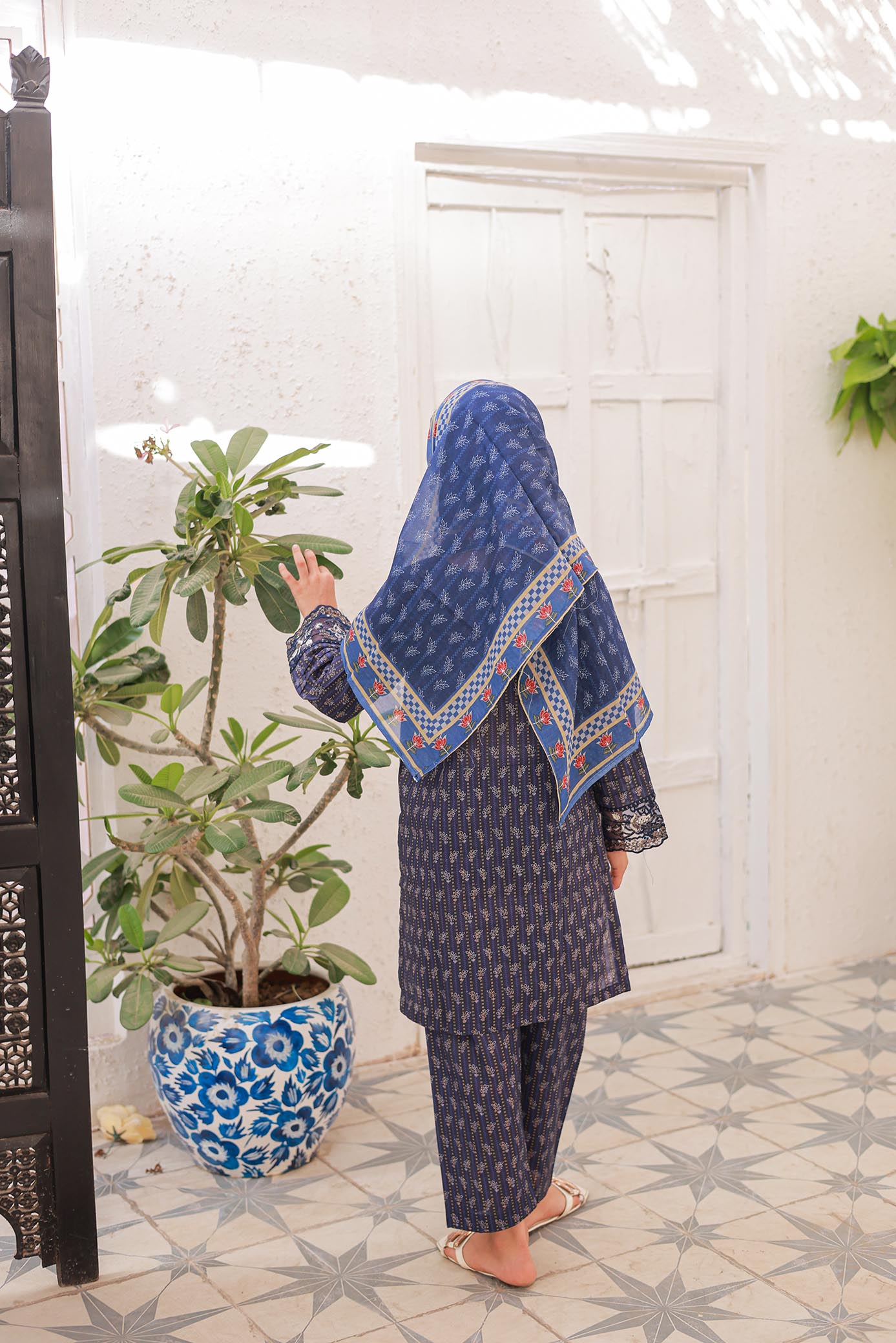 Person wearing a blue patterned hijab and outfit standing in a room with a plant and decorative items.