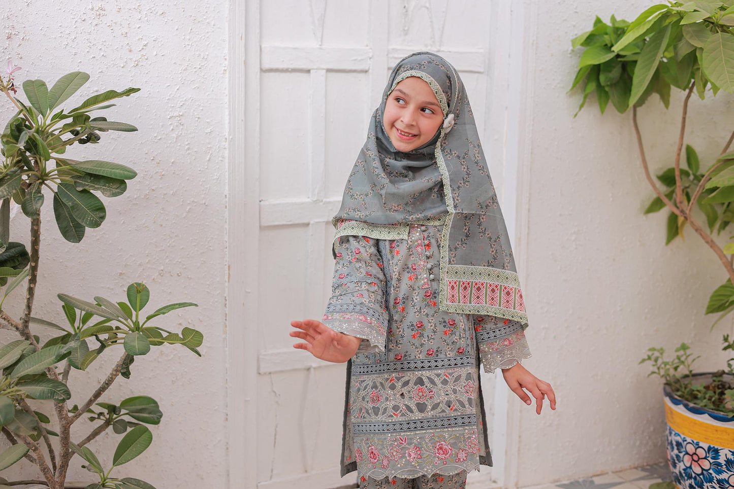 Young girl in a floral dress and hijab standing indoors with plants around.