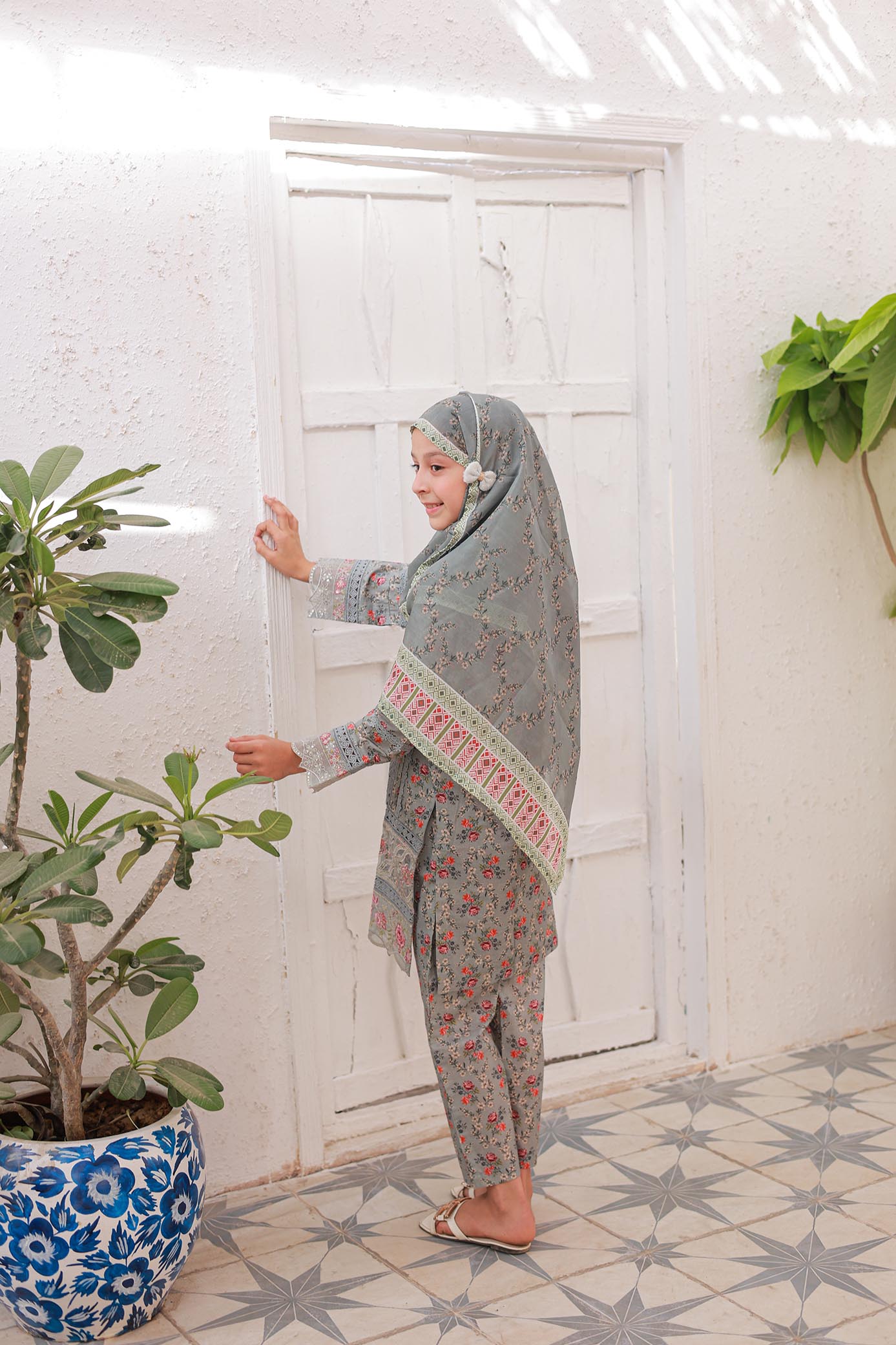 Woman in a patterned outfit standing in a room with plants and a white door.