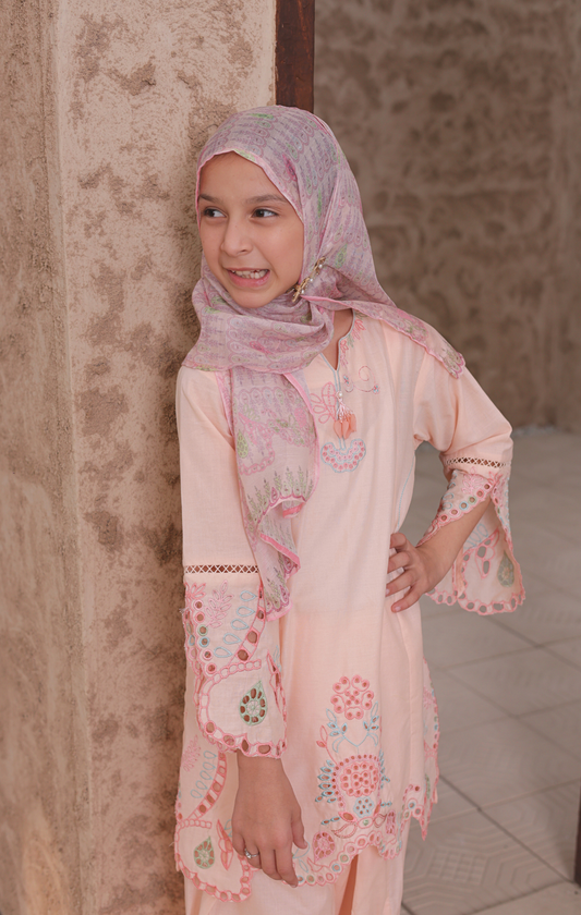 Young girl wearing a pink floral dress and hijab against a textured wall.