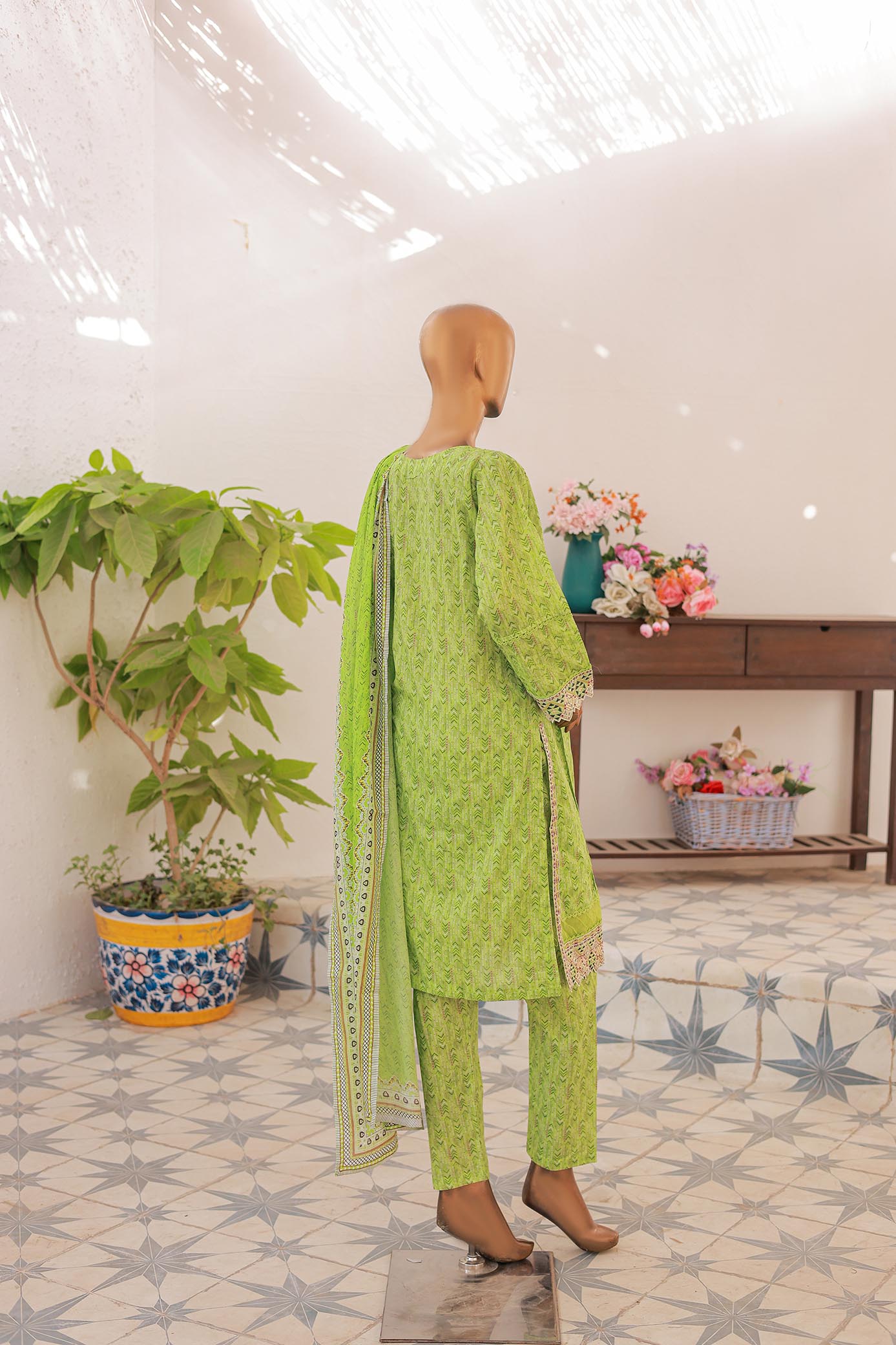 Mannequin wearing a green traditional outfit with a patterned dupatta in a room with plants and flowers.
