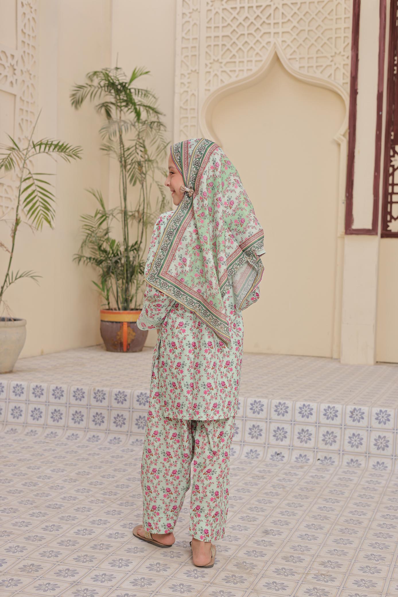 Woman in a floral outfit standing in a room with plants and decorative tiles.