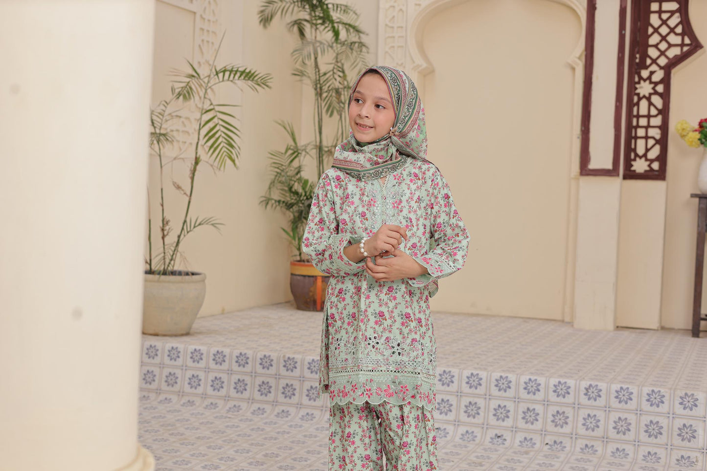 Young girl in a floral dress and headscarf standing in a decorated room with plants and a bed.