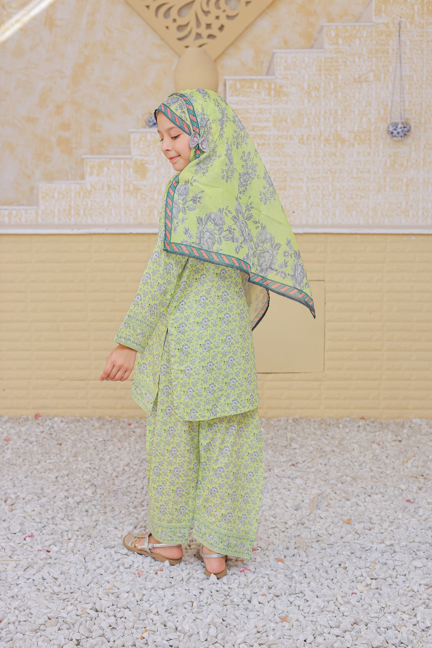 Woman in a green traditional outfit standing outdoors with a decorative wall in the background.