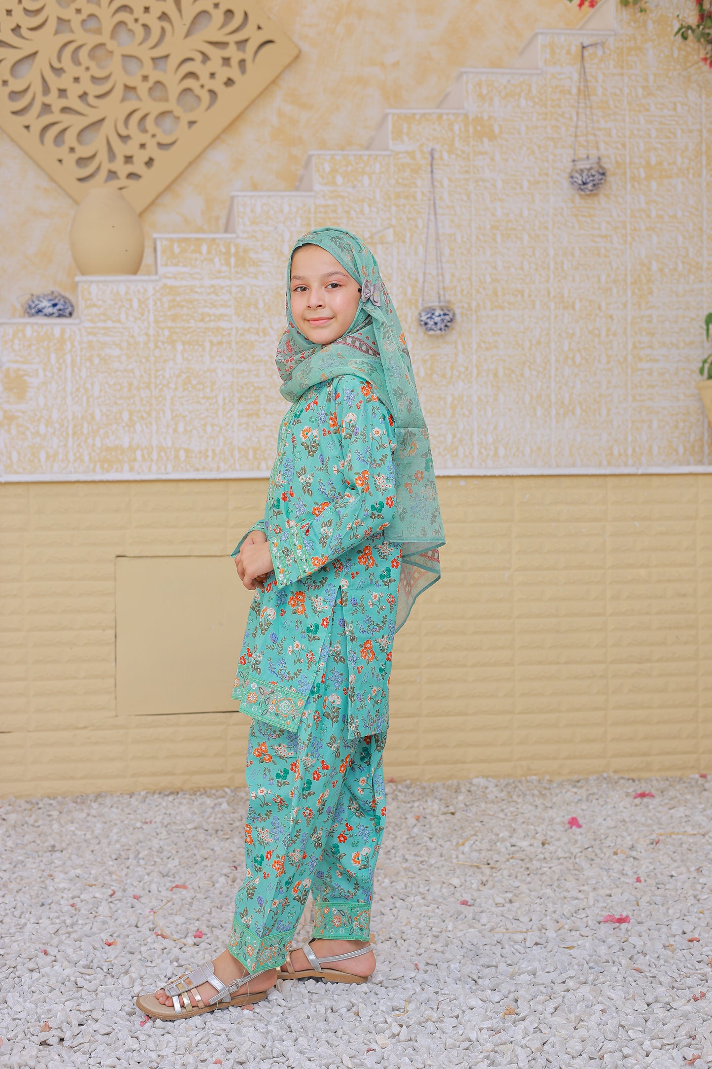 Child wearing a green traditional outfit with a floral pattern against a beige wall.