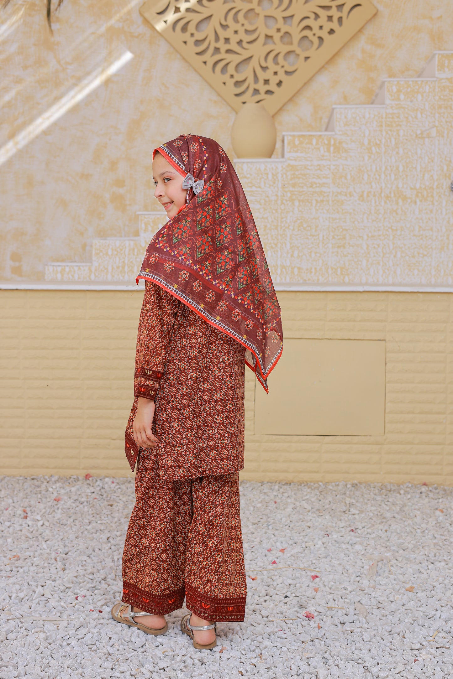 Woman wearing a traditional brown outfit with a matching headscarf in front of a decorative wall.