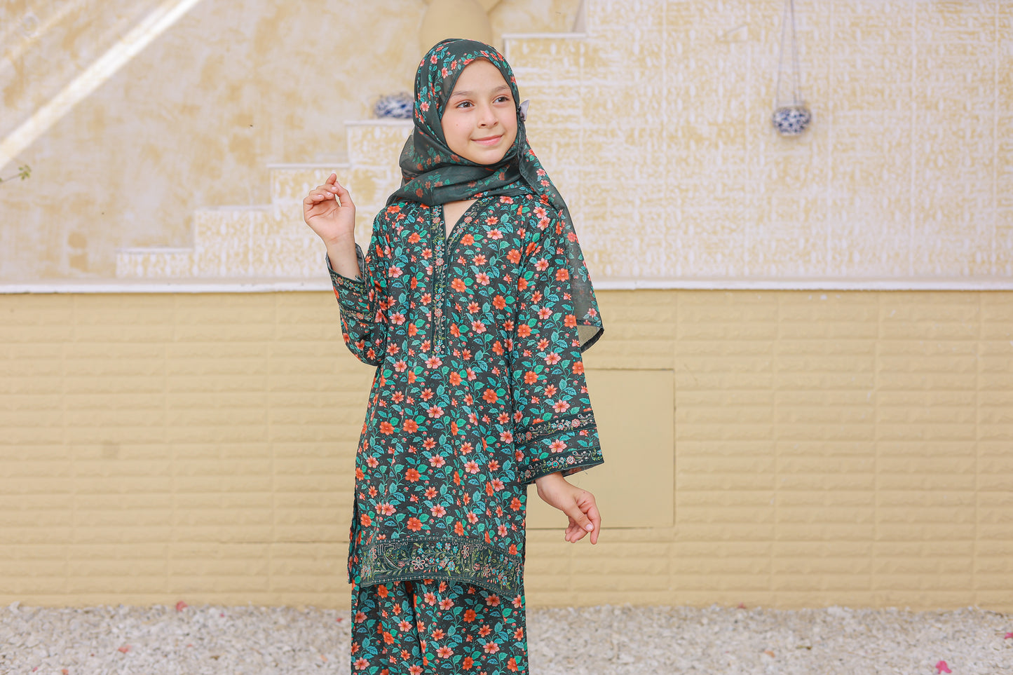 Child wearing a floral outfit with a headscarf standing against a beige wall.