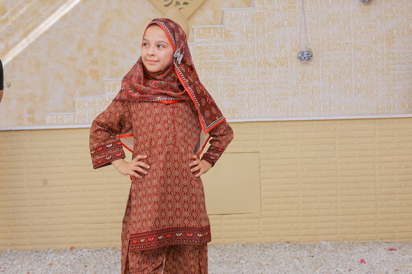 Young girl wearing a traditional red and patterned outfit with a headscarf in an indoor setting.