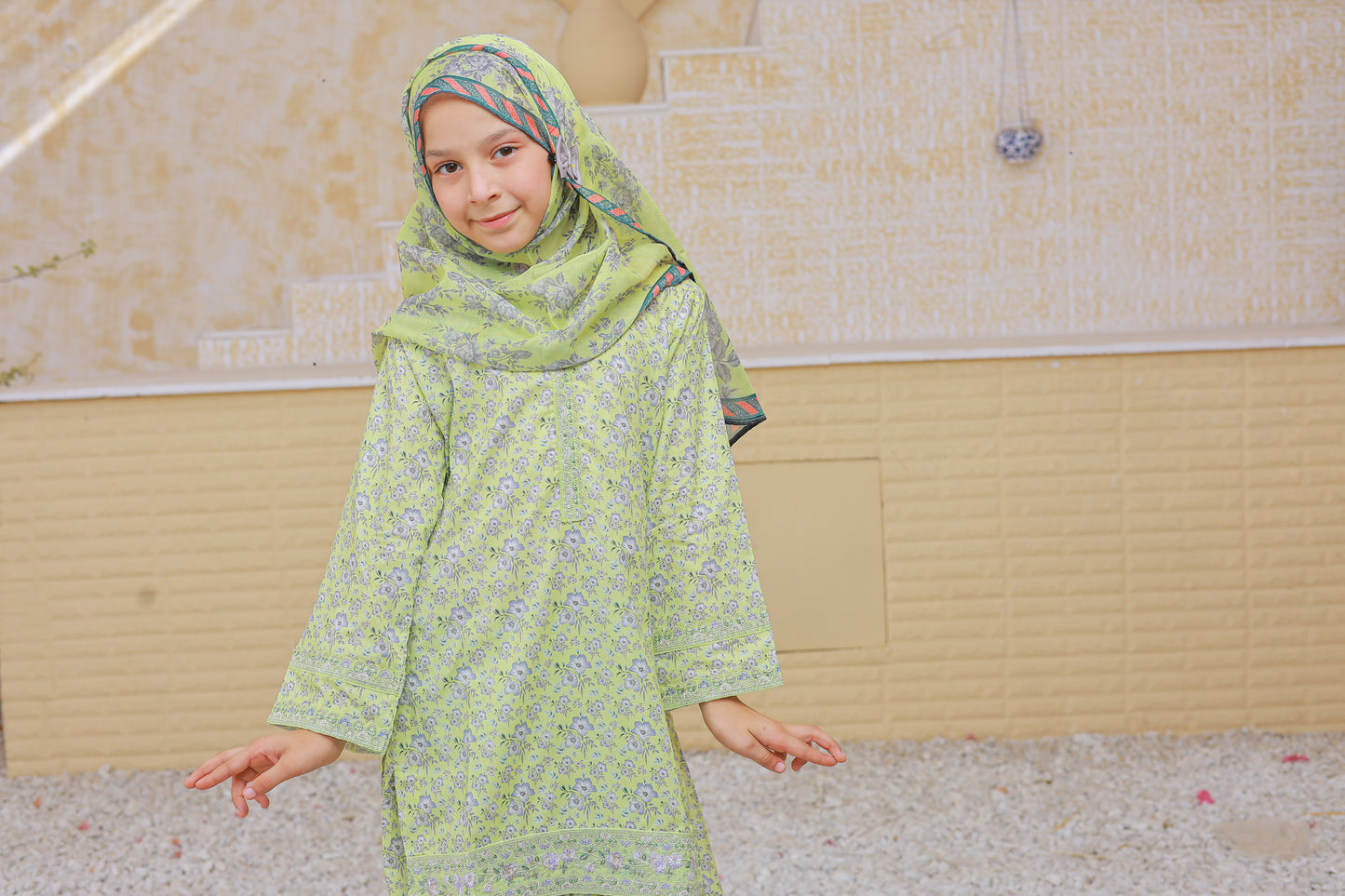 Young girl wearing a green traditional outfit with a headscarf in an indoor setting.