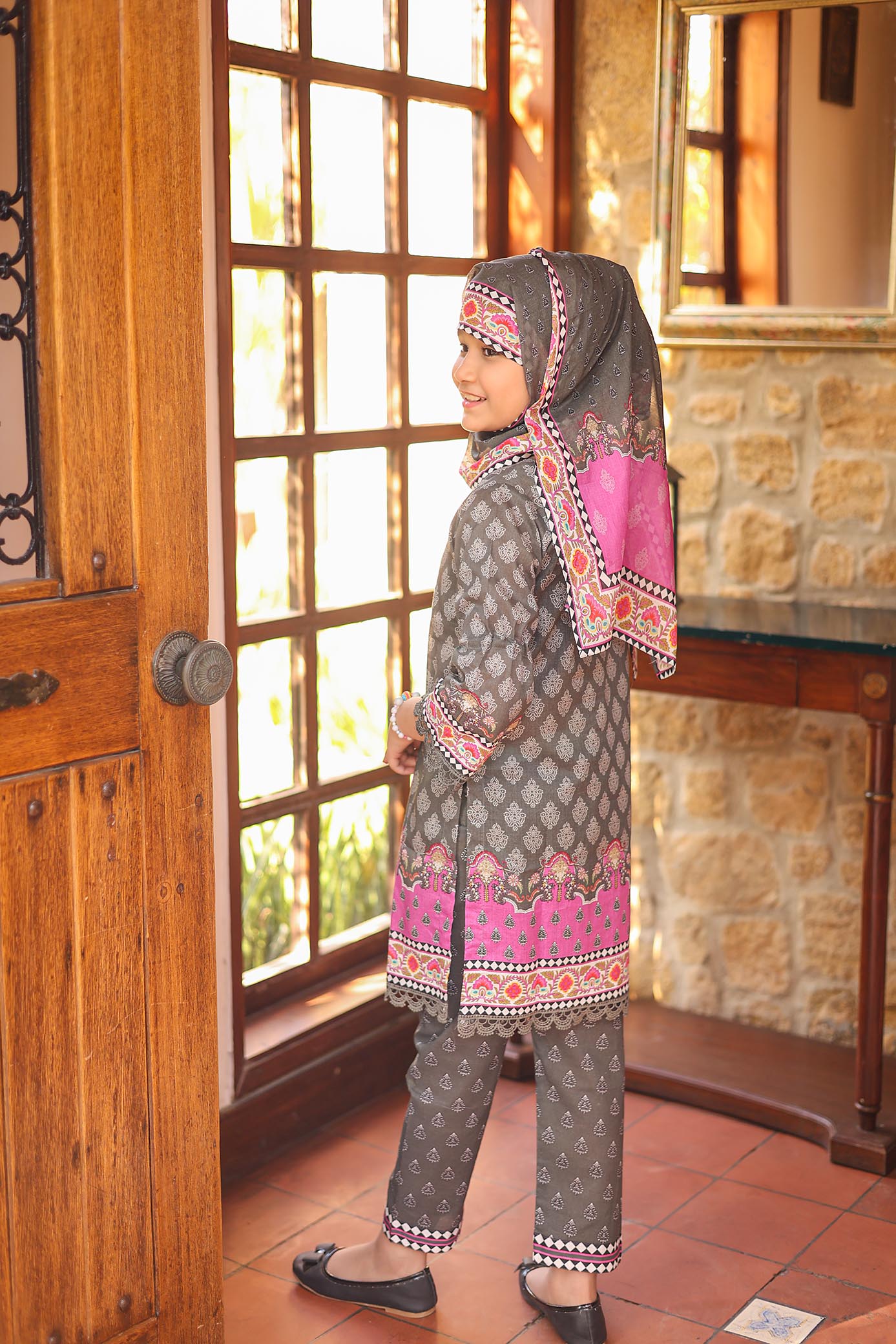 Woman in traditional attire standing in a room with wooden door and stone wall.
