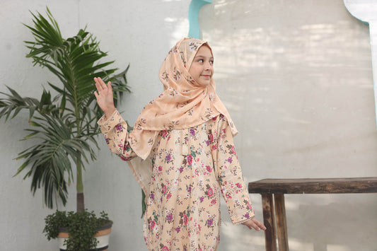Woman wearing a floral dress and hijab in an indoor setting with plants and a table.