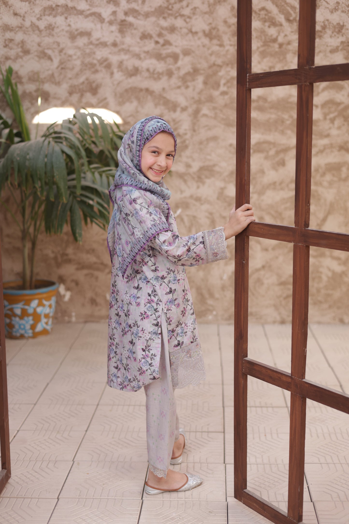 Woman in a floral dress and headscarf standing on a tiled floor with a plant in the background