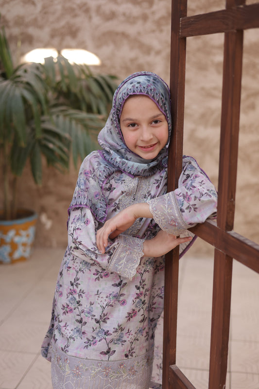 Woman in floral dress and hijab standing indoors with a plant in the background
