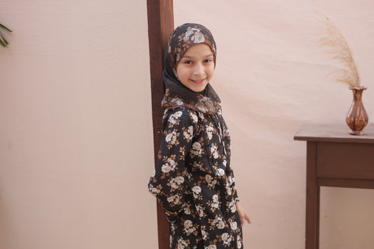 Young girl wearing a floral dress and hijab standing indoors against a neutral wall.