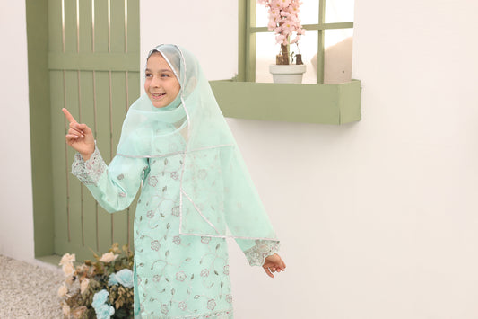 Young girl in a light green traditional outfit with a matching hijab, pointing towards the camera.