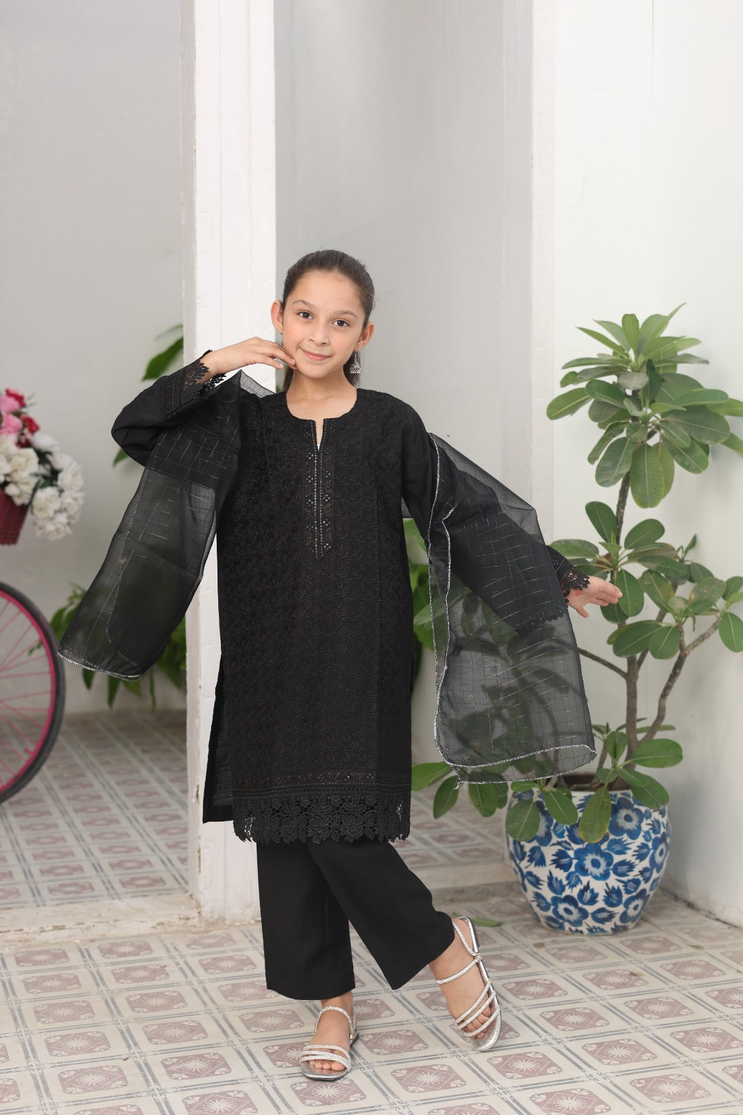 Young girl in a black outfit standing indoors with plants and a bicycle in the background