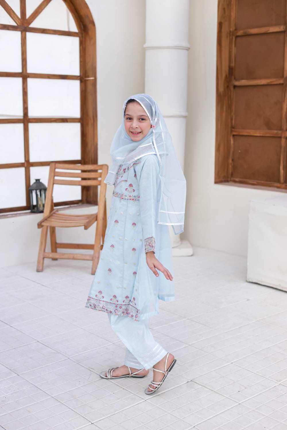 Woman in a light blue traditional outfit with a matching headscarf standing in a bright room.