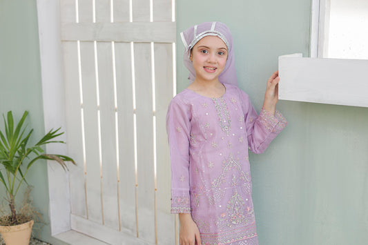 Young girl in a purple dress with floral patterns standing indoors against a light green wall.