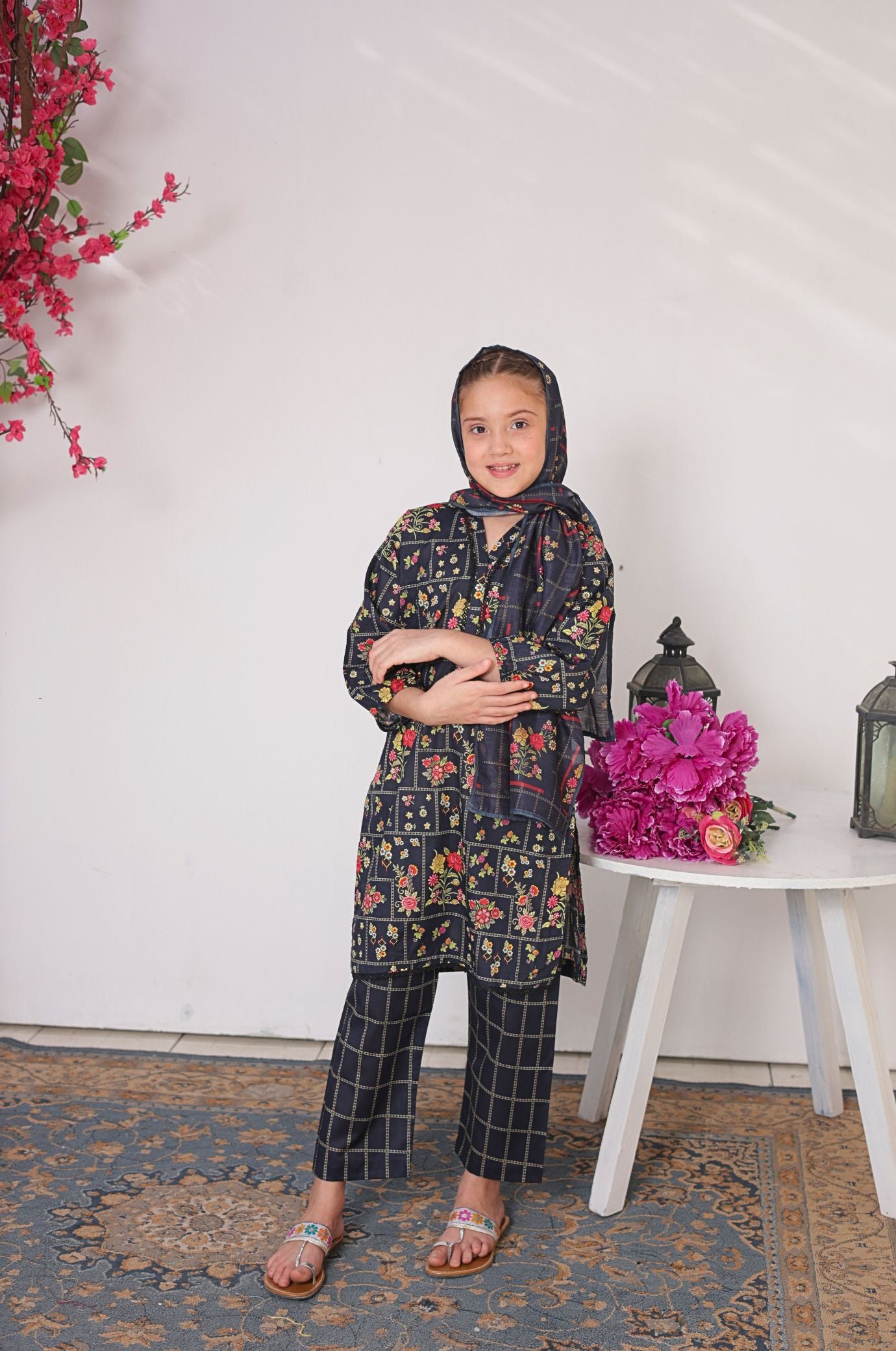 Young girl in a floral dress and headscarf standing next to a table with flowers and lanterns.