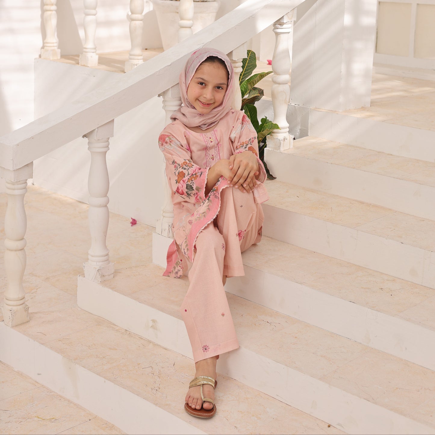 Woman in a pink dress sitting on a staircase with white railings and decorative plants.