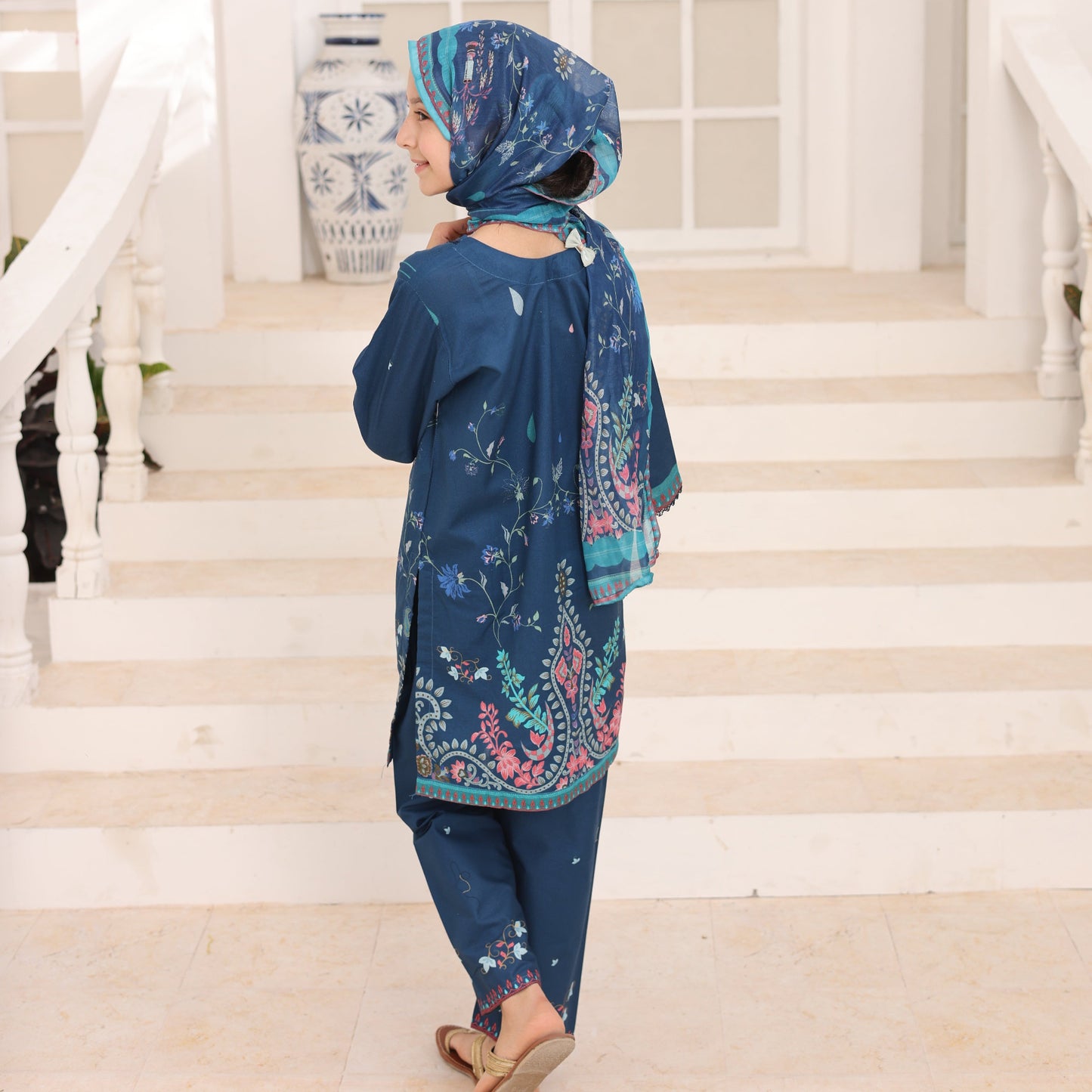 Woman wearing a blue dress with floral patterns in a room with white walls and a window.