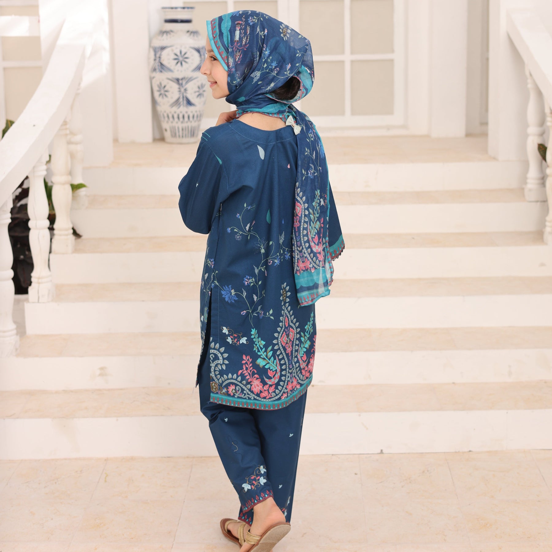 Woman wearing a blue dress with floral patterns in a room with white walls and a window.