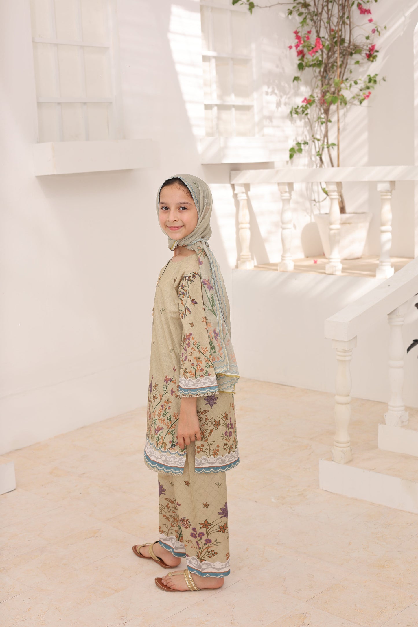 Woman in a floral dress and hijab standing in a room with a mirror and decorative plants.