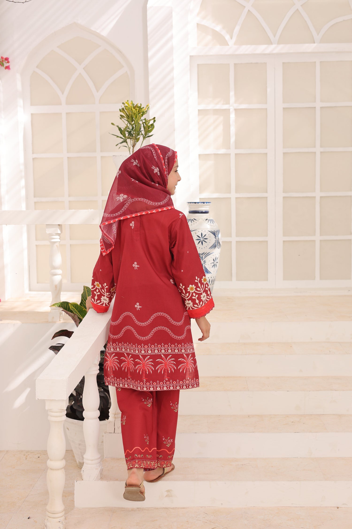 Woman in a red traditional outfit standing on a staircase with a decorative wall in the background.