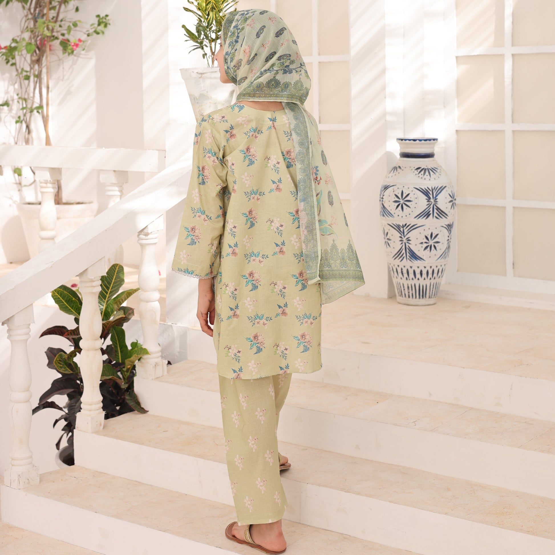 Person wearing a light green floral outfit standing in a room with decorative plants and a vase.
