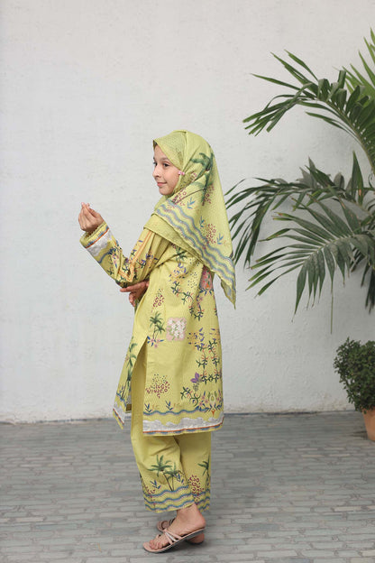 Child wearing a green traditional outfit with a matching headscarf, standing against a white wall with plants.