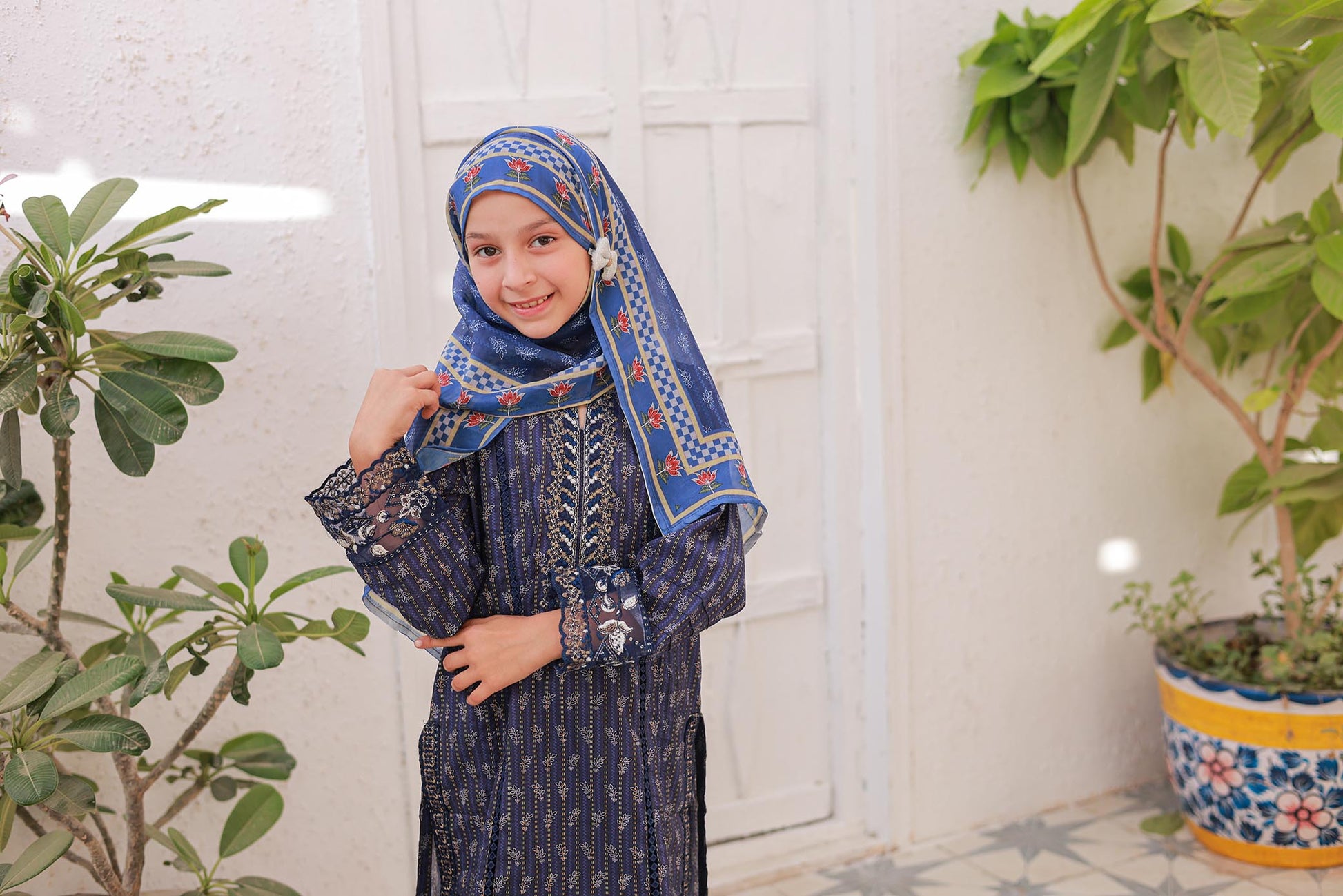 Child wearing a blue and white patterned hijab and dress, standing indoors with plants in the background.