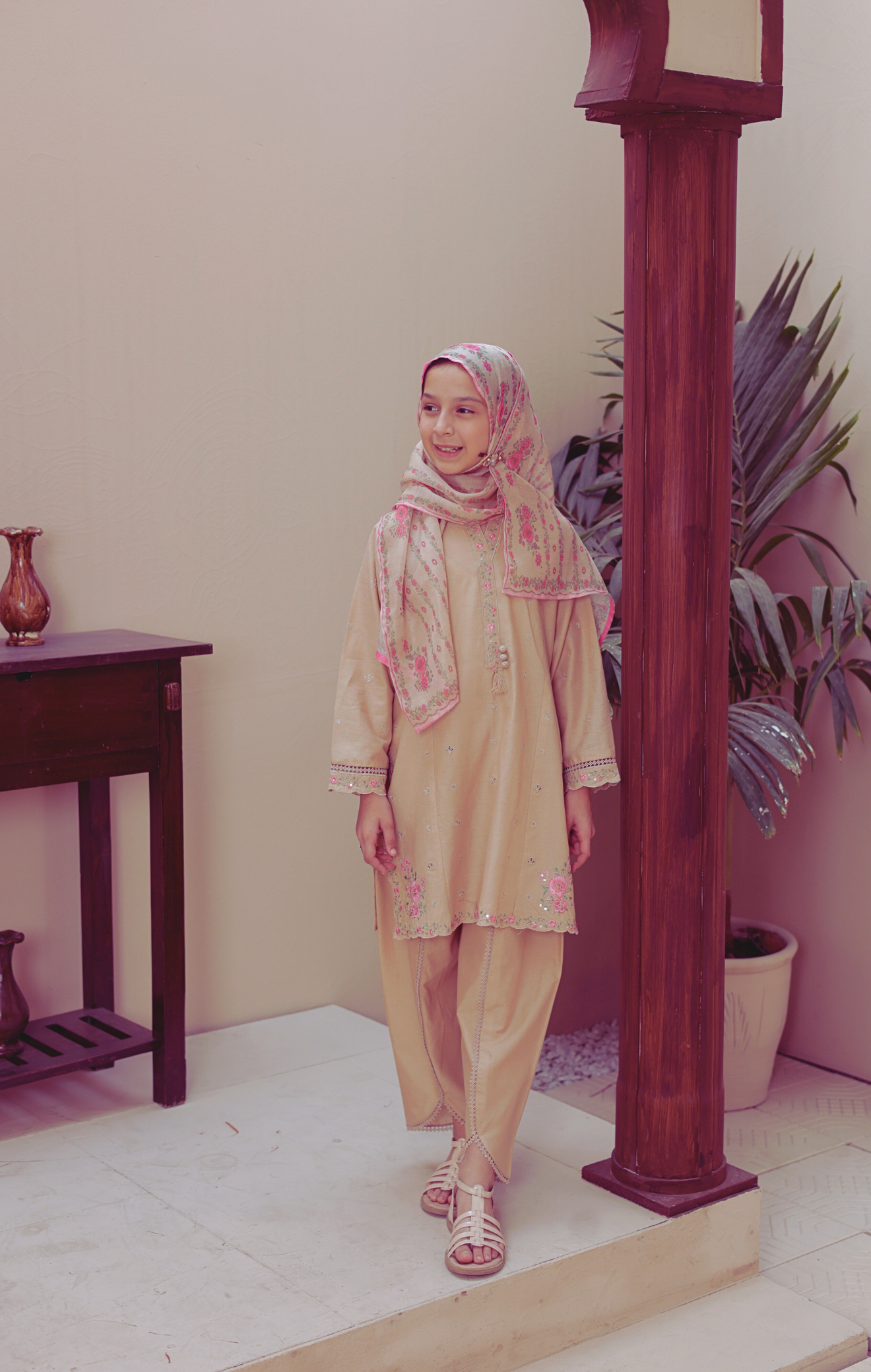 Woman in a beige outfit with a floral headscarf standing indoors next to a wooden column.