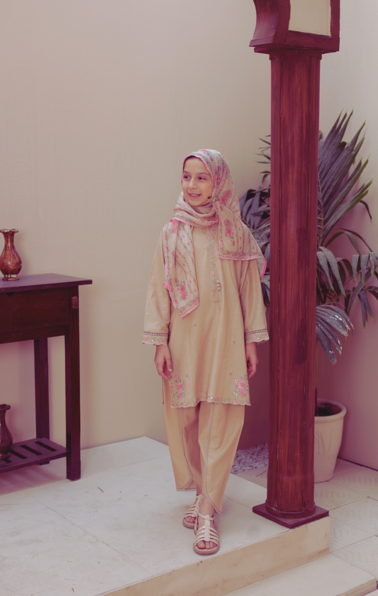 Woman in a beige outfit with a floral headscarf standing indoors next to a wooden column.