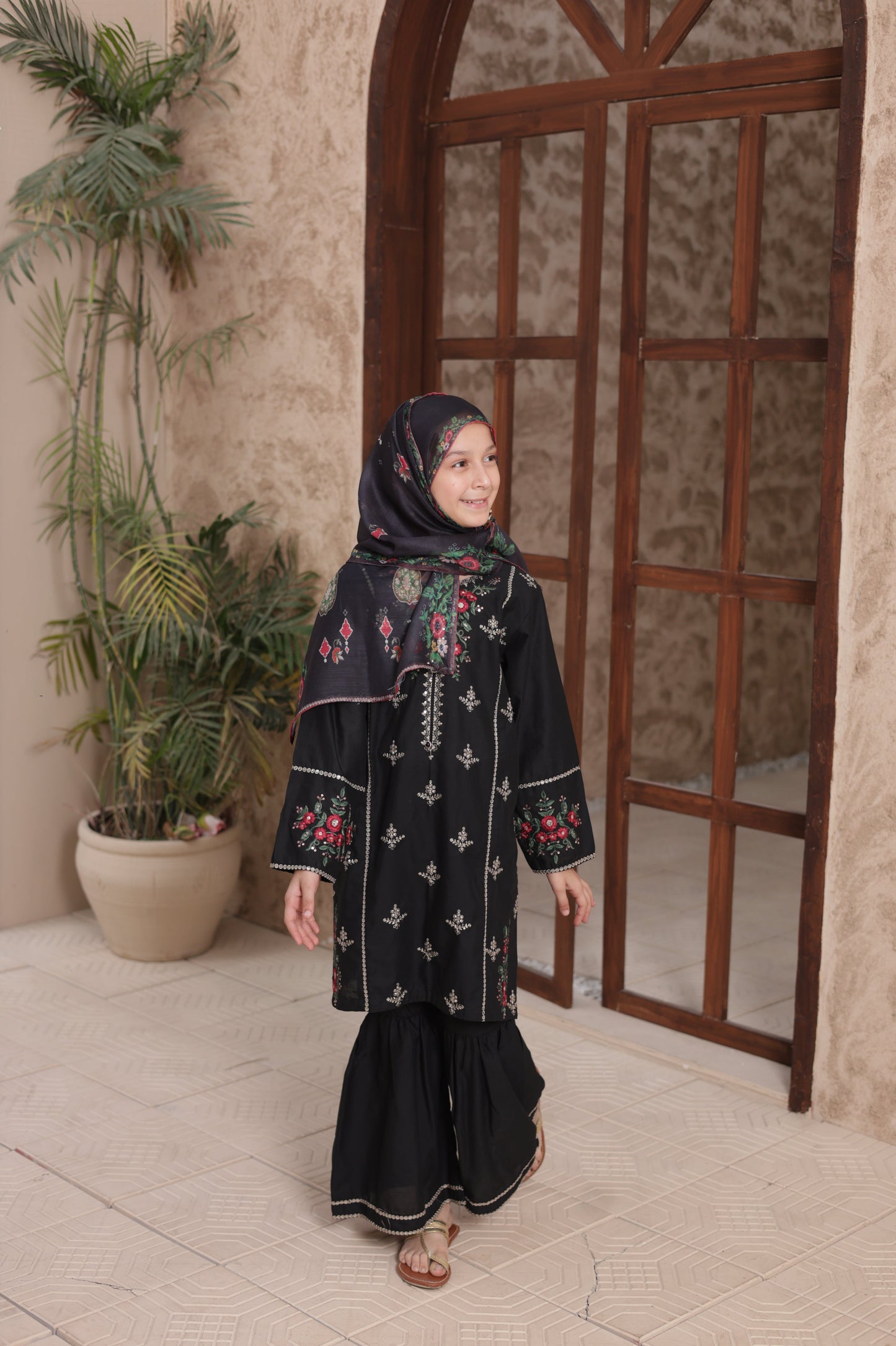 Person wearing a black embroidered dress standing in a room with a wooden chair and plants.