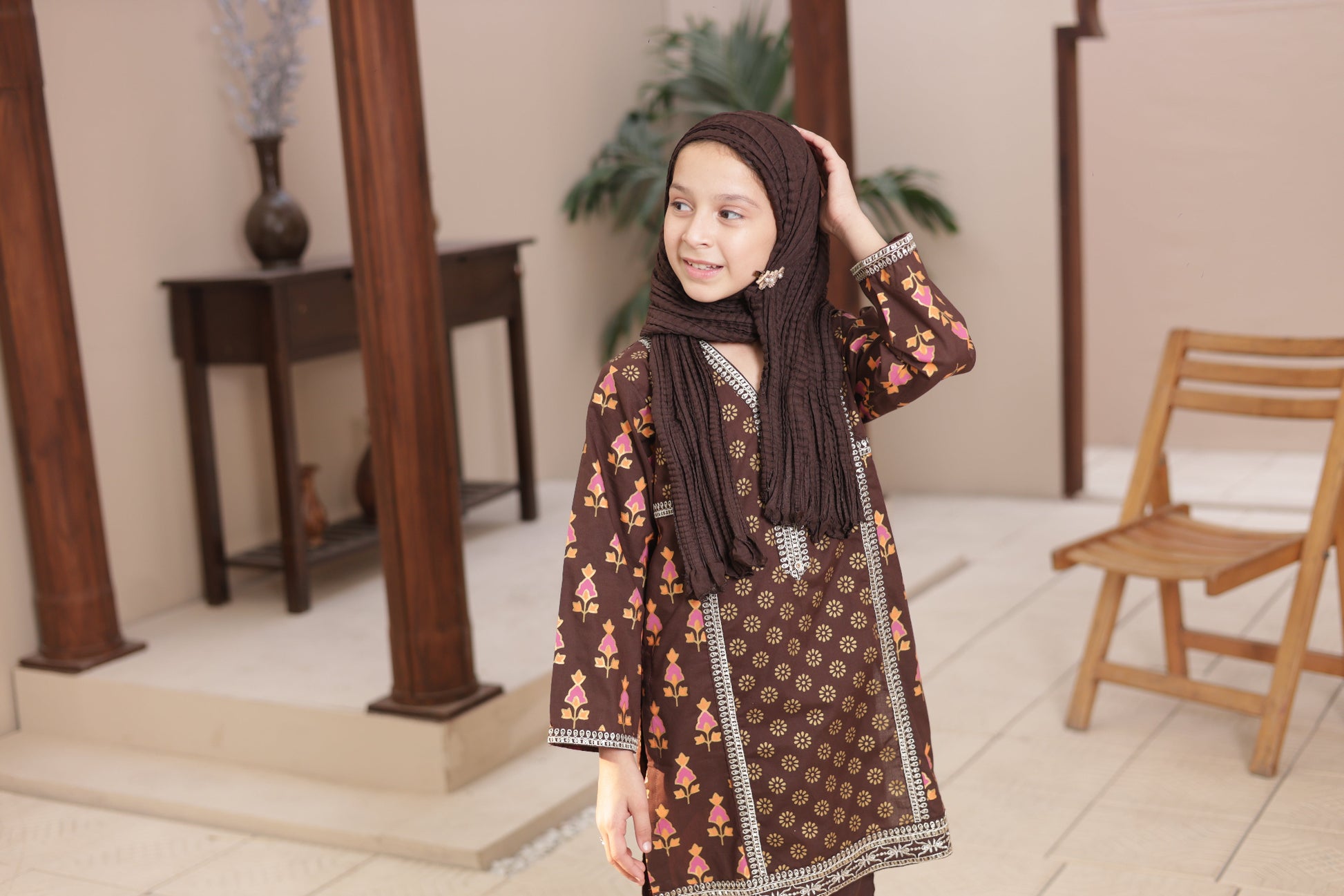 Young girl wearing a patterned dress and hijab indoors.