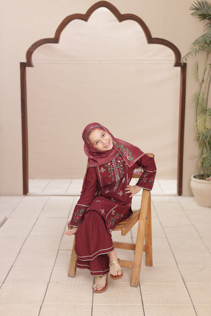 Child in a red outfit with floral patterns sitting on a wooden stool against a white wall.
