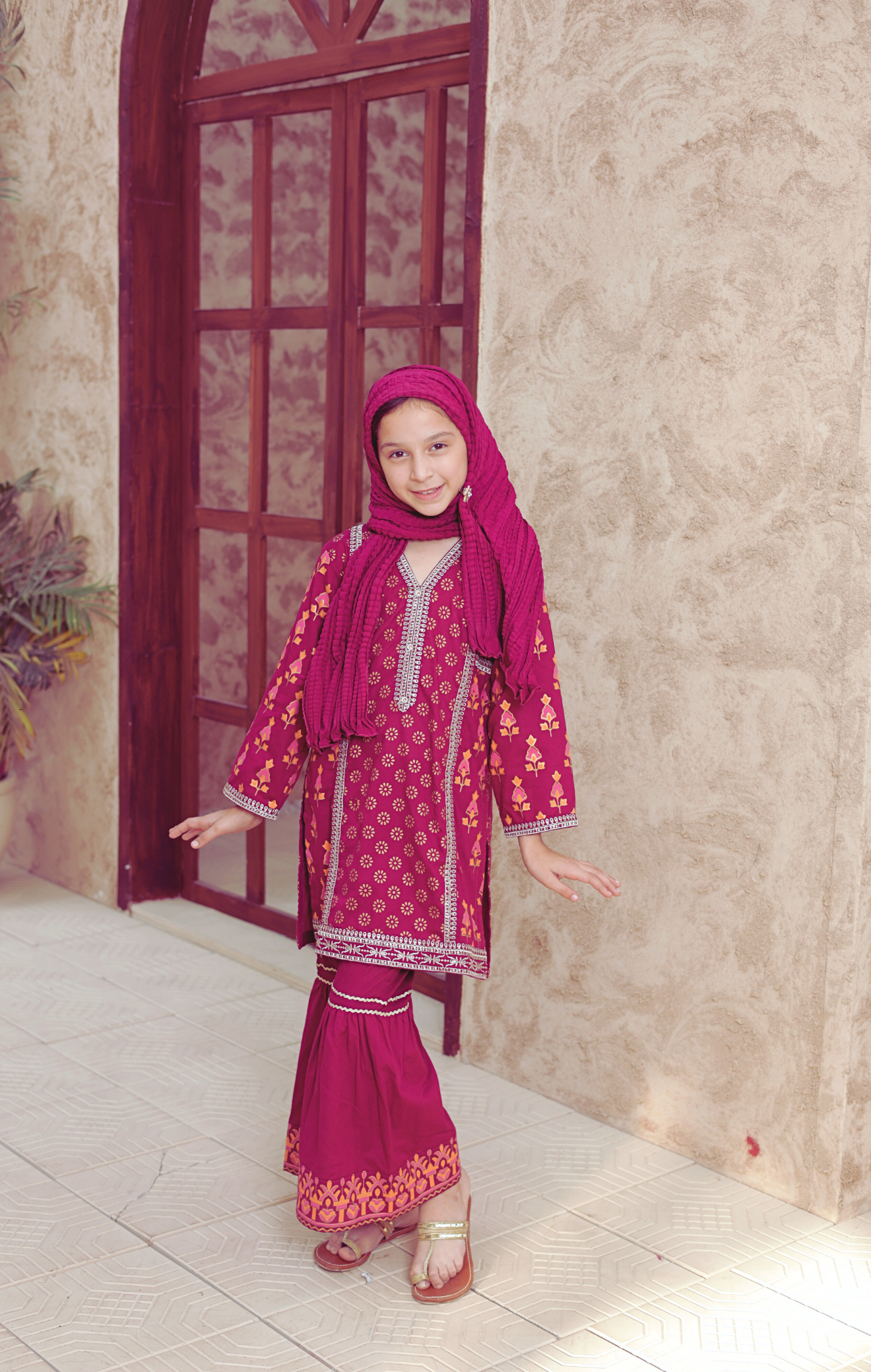 Young girl in a traditional outfit with a pink headscarf standing in a room with a red door and beige walls.