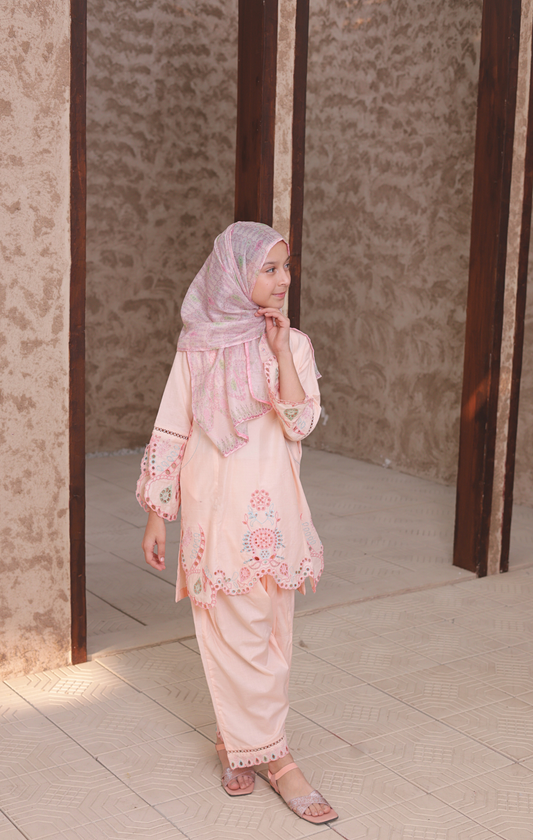 Woman in a light pink traditional outfit with floral embroidery standing in a doorway.