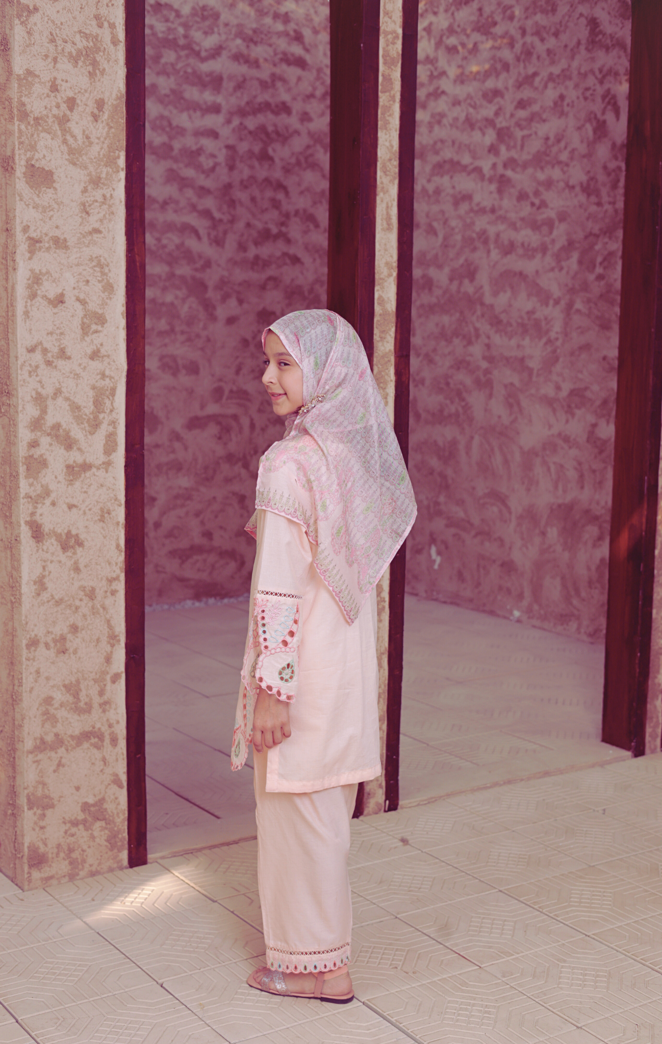 Young girl in a pink traditional outfit standing in a room with patterned walls.