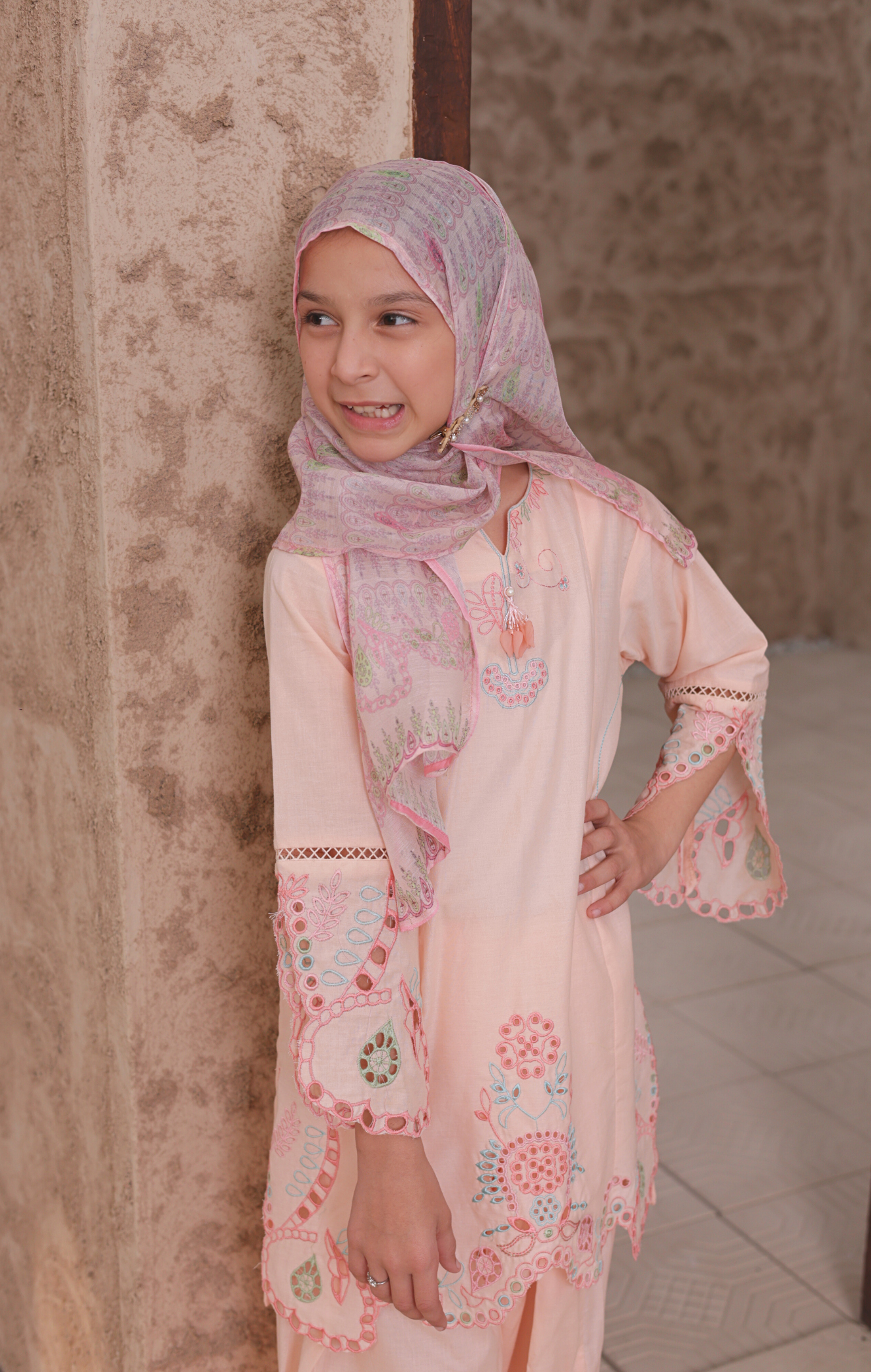 Young girl wearing a pink floral dress and hijab against a textured wall.