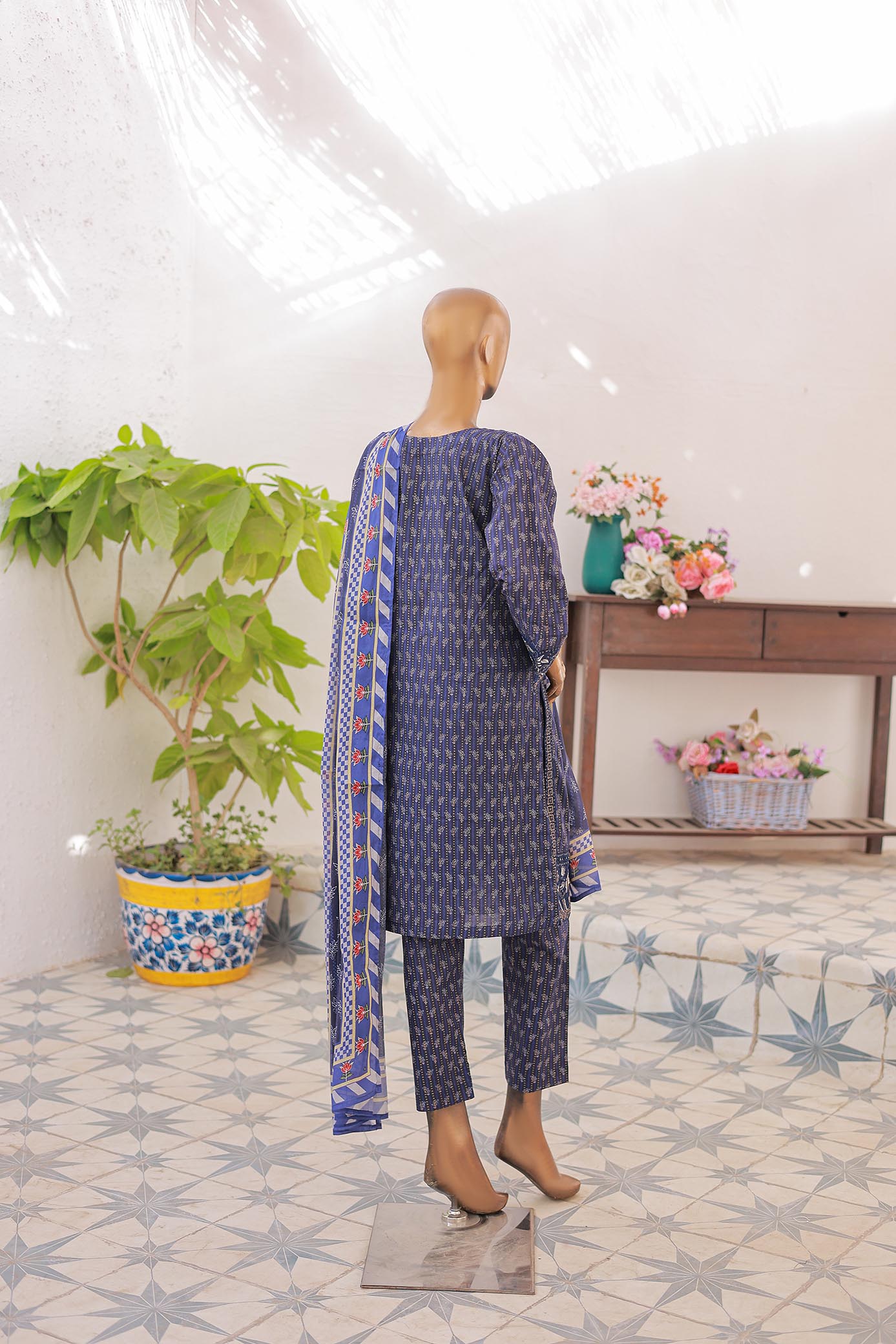 Mannequin wearing a blue patterned outfit with a scarf in a room with plants and flowers.