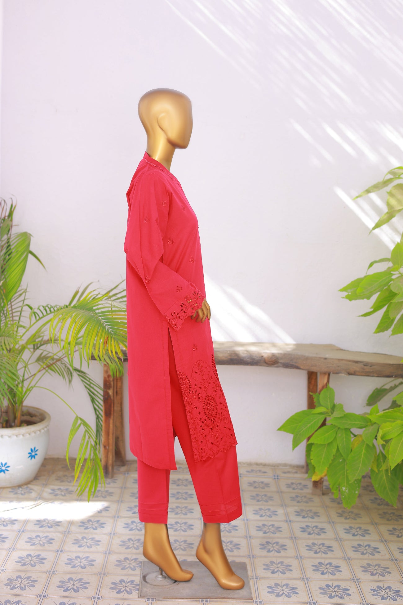 Mannequin wearing a red outfit with lace details in a room with plants and a bench.
