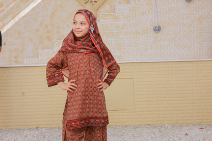 Young girl wearing a traditional red and patterned outfit with a headscarf in an indoor setting.