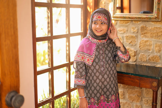 Young girl wearing a patterned dress and headscarf standing in front of a stone wall and window.